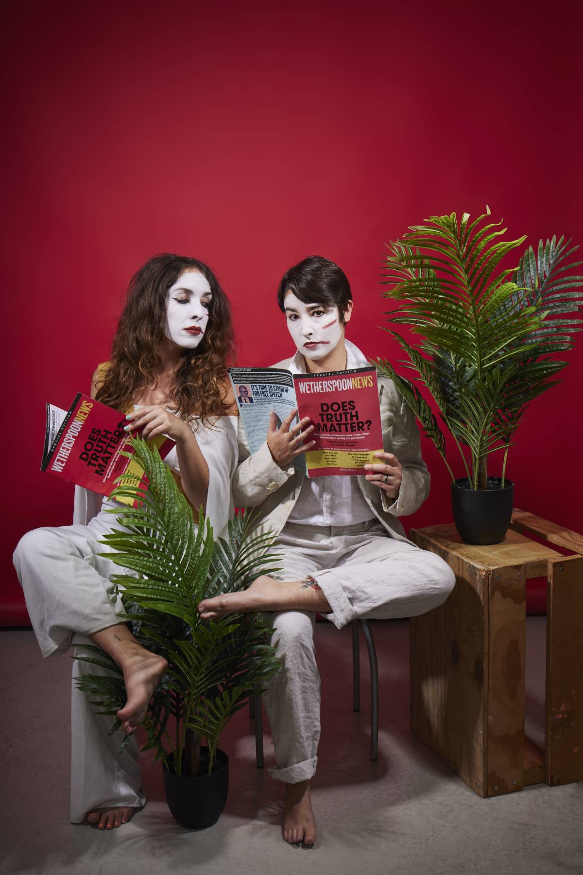 two women in white face paint reading magazines sat amongst office plants