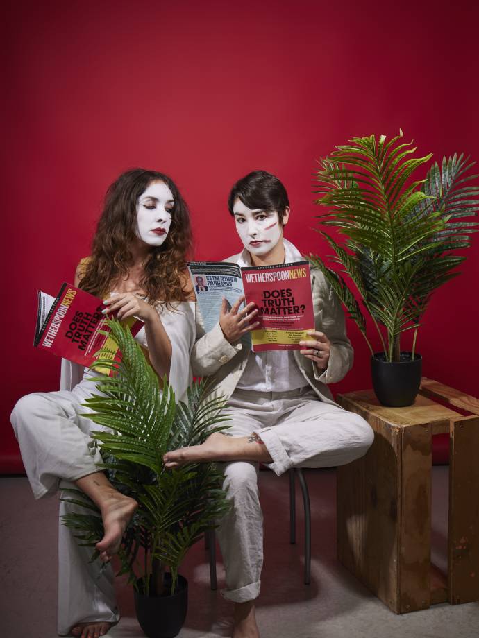 two women in white face paint reading magazines sat amongst office plants