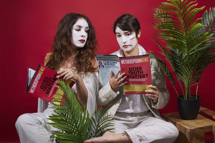 two women in white face paint reading magazines sat amongst office plants