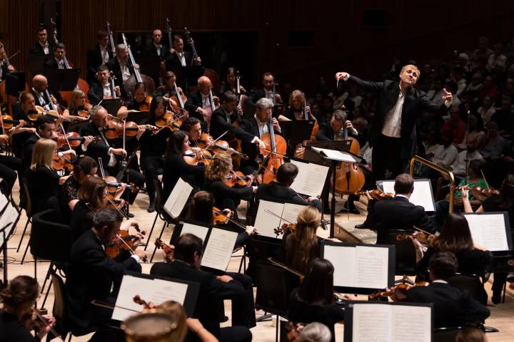 Conductor in a white shirt and dark jacket leading a large, seated orchestra of formally dressed musicians on a wooden stage.